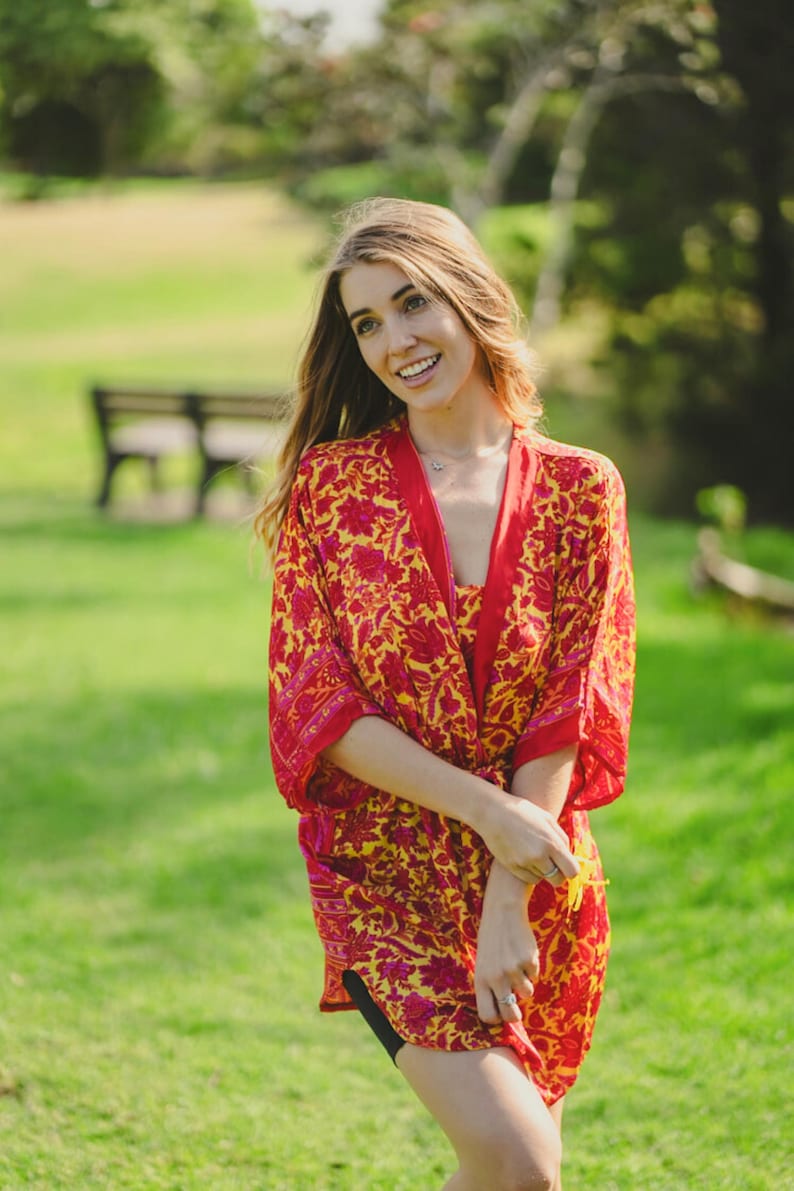 May include: A vibrant, knee-length kimono-style robe with a floral pattern in red and yellow. The robe has a contrasting red trim and is worn outdoors on a sunny day. The garment is lightweight and appears to be made of a flowing fabric.