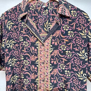 May include: Black button-down shirt with a floral pattern in pink and gold. The shirt has short sleeves and a collared neckline. The center placket and collar edges feature a pink and gold stripe design.