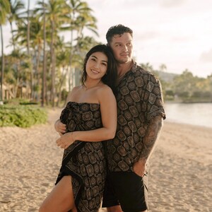 Puede incluir: Una pareja en una playa, vistiendo ropa a juego con estampado negro y dorado. La mujer lleva un pareo sin tirantes y el hombre una camisa abotonada y pantalones cortos negros. Palmeras y una playa de arena en el fondo.