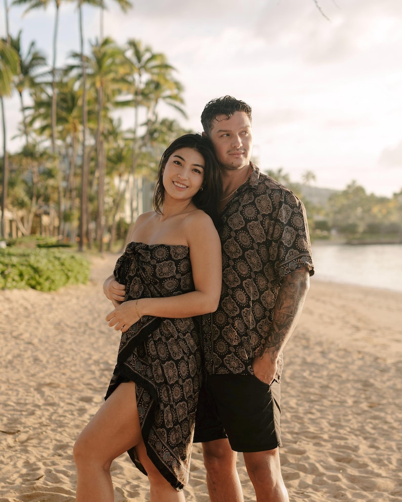 Puede incluir: Una pareja en una playa, vistiendo ropa a juego con estampado negro y dorado. La mujer lleva un pareo sin tirantes y el hombre una camisa abotonada y pantalones cortos negros. Palmeras y una playa de arena en el fondo.