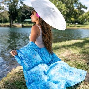 May include: A bright blue shawl featuring a detailed paisley pattern in shades of light and dark blue, draped over a beige tank top. The shawl is paired with a plain white paper parasol held overhead, creating a contrast between the vibrant fabric.