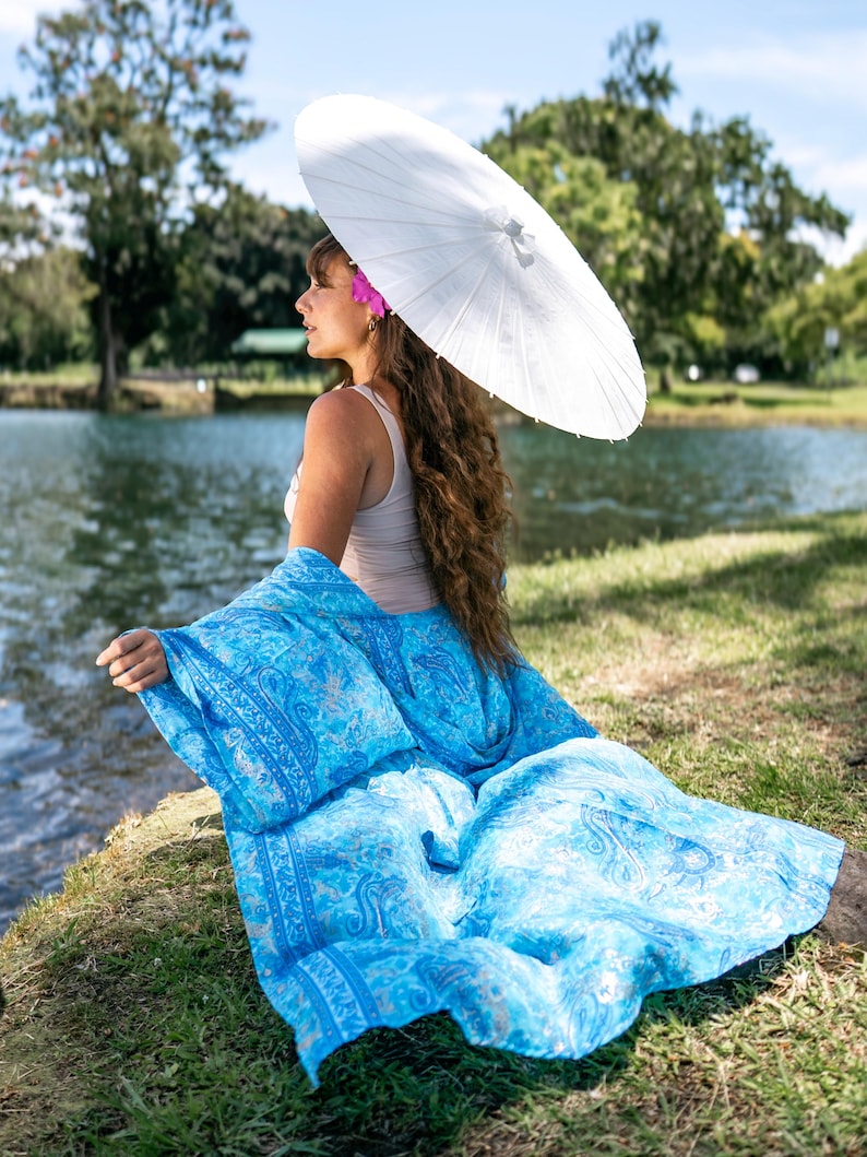 May include: A bright blue shawl featuring a detailed paisley pattern in shades of light and dark blue, draped over a beige tank top. The shawl is paired with a plain white paper parasol held overhead, creating a contrast between the vibrant fabric.