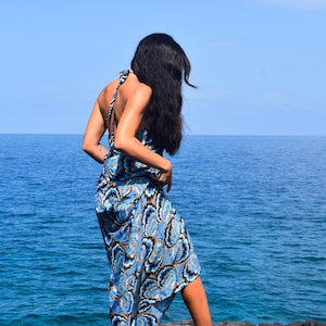 May include: A blue and white patterned halter dress with a flowing skirt. The dress is worn by a person standing on a rocky surface, with the ocean and blue sky in the background.