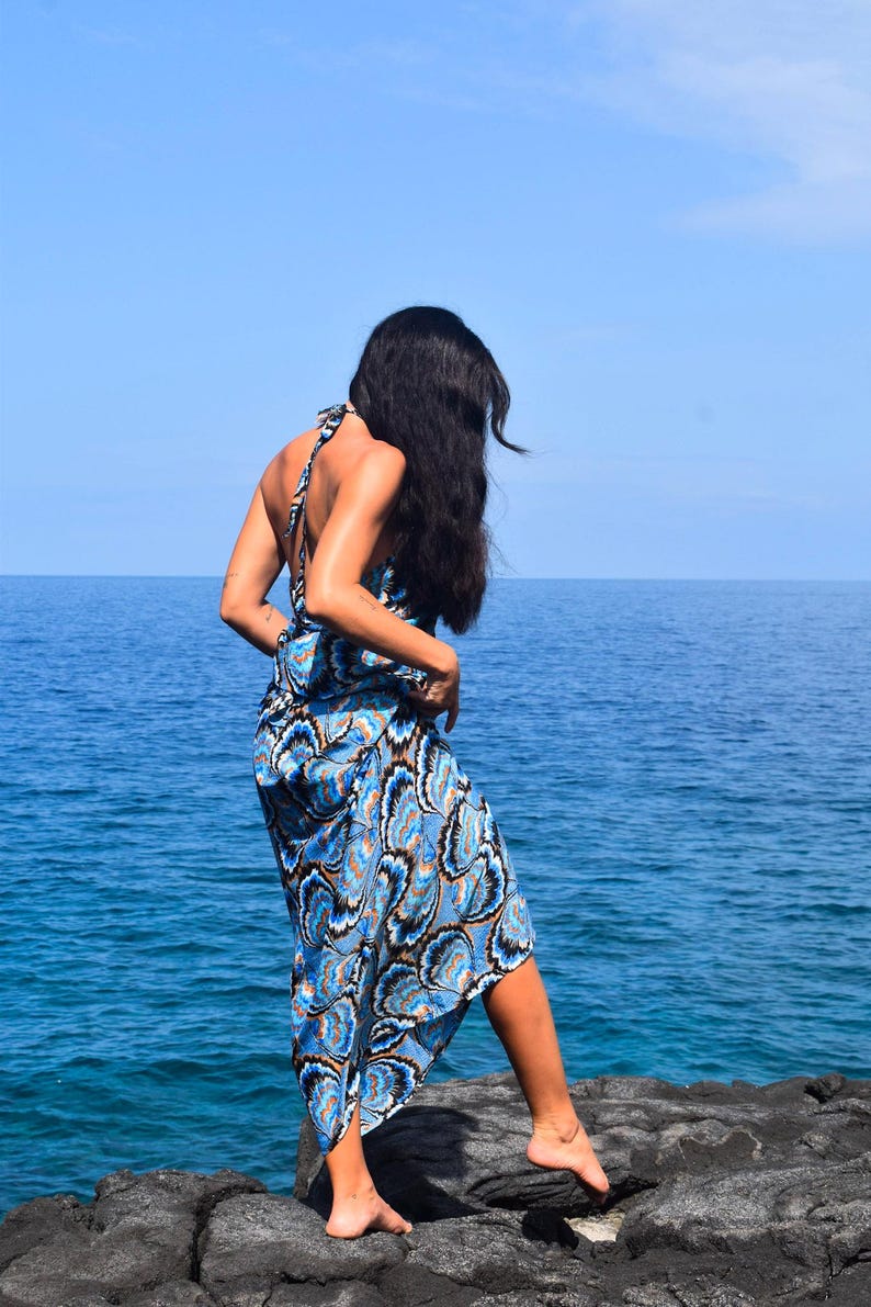 May include: A blue and white patterned halter dress with a flowing skirt. The dress is worn by a person standing on a rocky surface, with the ocean and blue sky in the background.