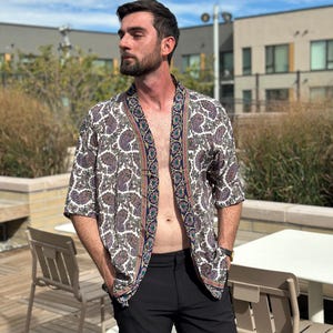 May include: A short-sleeved paisley print kimono in white, brown, and purple with a navy blue and green border. The kimono is paired with black shorts. The image is taken outdoors on a sunny day.