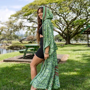 Puede incluir: Una bata con capucha verde con un estampado floral. La bata tiene un dise&ntilde;o largo y fluido y est&aacute; hecha de una tela ligera. El estampado presenta tonos de verde y azul con un toque de rojo. La bata se usa al aire libre en un parque.