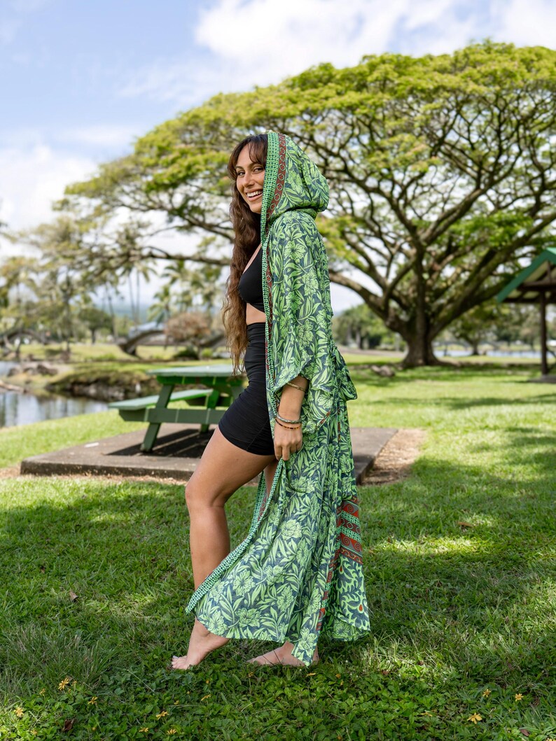 Puede incluir: Una bata con capucha verde con un estampado floral. La bata tiene un dise&ntilde;o largo y fluido y est&aacute; hecha de una tela ligera. El estampado presenta tonos de verde y azul con un toque de rojo. La bata se usa al aire libre en un parque.