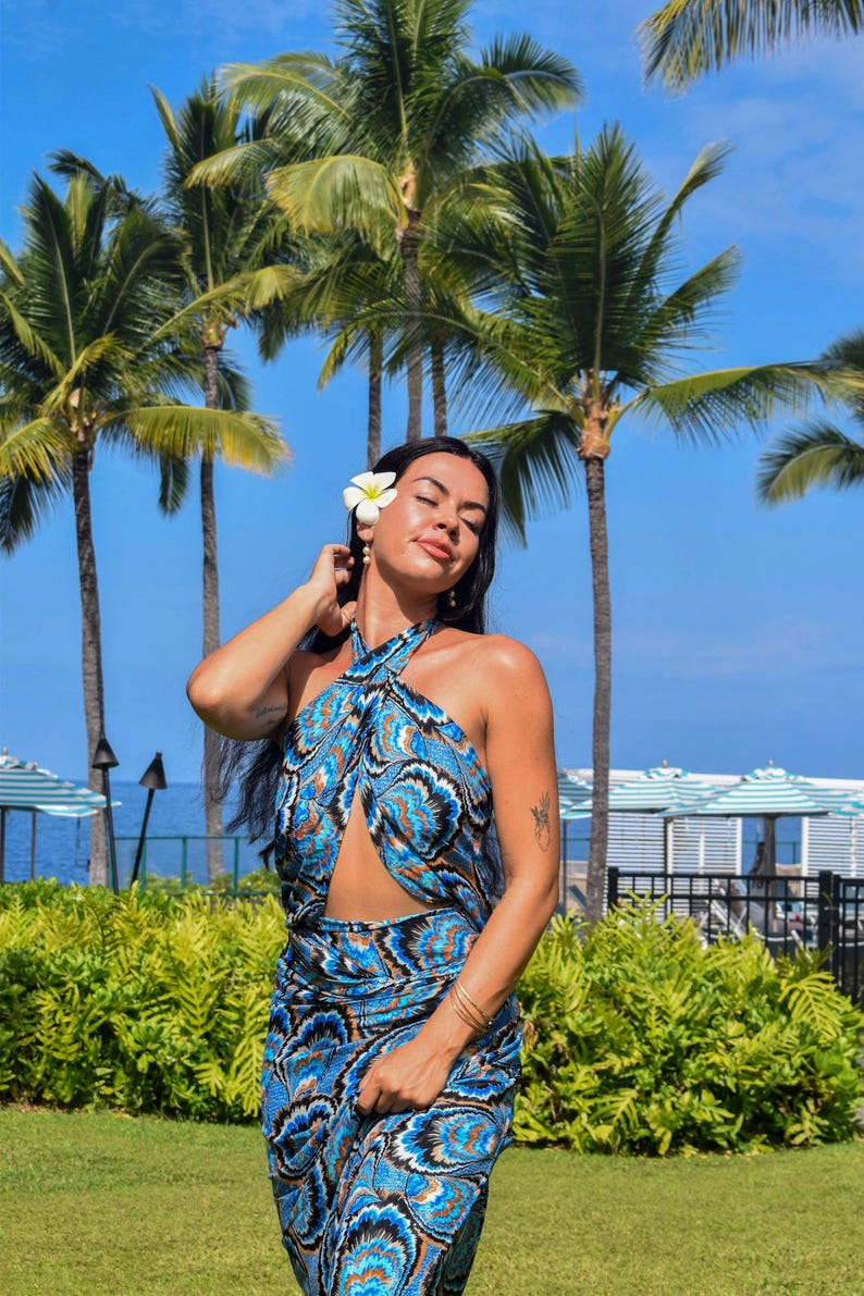 May include: A woman wearing a blue and turquoise patterned halter top and skirt, with a cut-out design. She has a white flower in her hair and is standing in front of palm trees and a blue sky. The outfit has a paisley-like design.