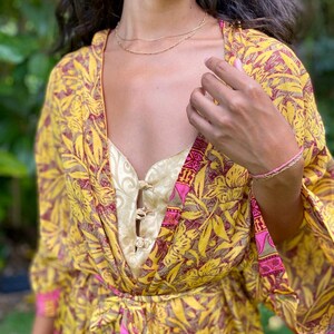 May include: A yellow and maroon floral print kimono robe with wide sleeves and a matching waist tie, worn over a pale gold sleeveless top featuring a front placket with three decorative toggle closures and a subtle swirling pattern.