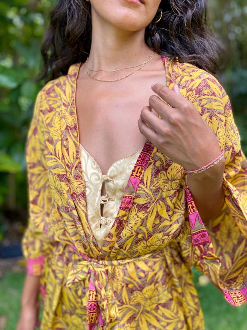 May include: A yellow and maroon floral print kimono robe with wide sleeves and a matching waist tie, worn over a pale gold sleeveless top featuring a front placket with three decorative toggle closures and a subtle swirling pattern.