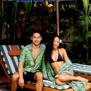 Puede incluir: Una pareja est&aacute; sentada en tumbonas de madera bajo una sombrilla azul y blanca. El hombre lleva una camisa verde con estampado de hojas y pantalones cortos azul marino. La mujer lleva un top de bikini negro y una envoltura verde estampada. Tema tropical.