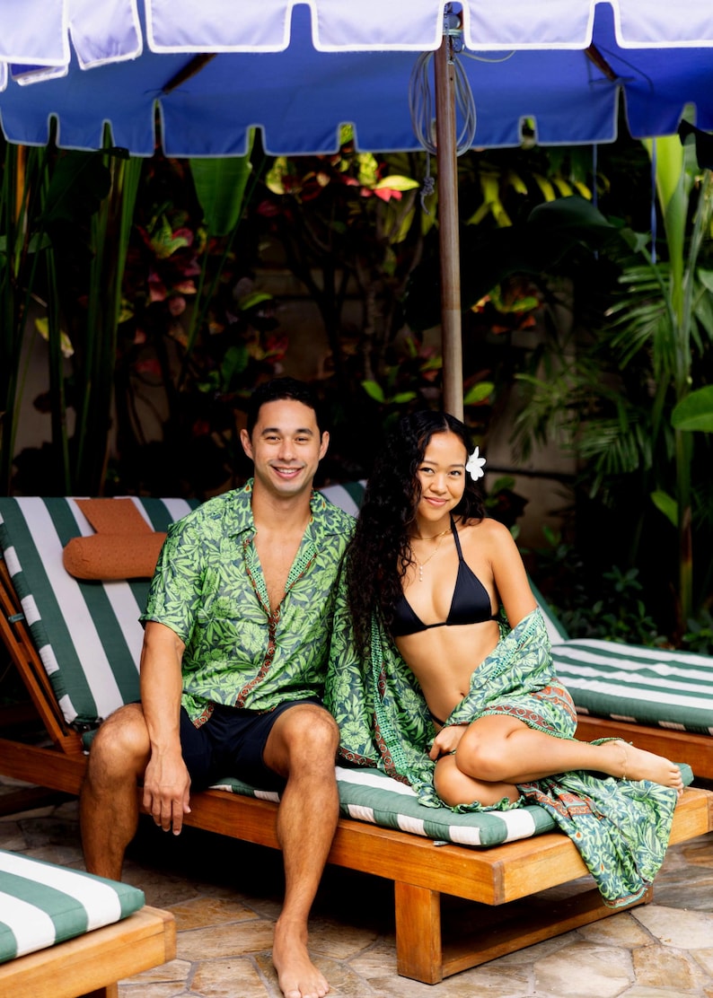 Puede incluir: Una pareja est&aacute; sentada en tumbonas de madera bajo una sombrilla azul y blanca. El hombre lleva una camisa verde con estampado de hojas y pantalones cortos azul marino. La mujer lleva un top de bikini negro y una envoltura verde estampada. Tema tropical.