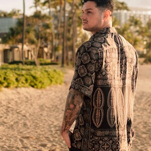 Puede incluir: Una camisa de manga corta con fondo negro y estampado de cachemira en tonos marr&oacute;n, beige y azul. La camisa presenta un dise&ntilde;o repetitivo de formas florales y geom&eacute;tricas. La camisa se lleva en una playa.