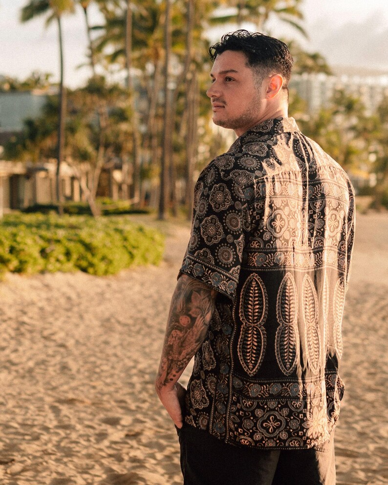 Puede incluir: Una camisa de manga corta con fondo negro y estampado de cachemira en tonos marr&oacute;n, beige y azul. La camisa presenta un dise&ntilde;o repetitivo de formas florales y geom&eacute;tricas. La camisa se lleva en una playa.