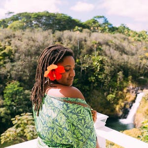 Puede incluir: Una mujer con rastas y una flor roja y naranja en el pelo, con una envoltura verde con estampado floral. El fondo presenta una cascada y un exuberante follaje verde, con una barandilla blanca en primer plano.
