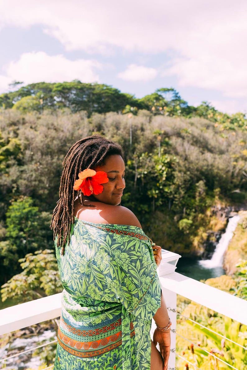 Puede incluir: Una mujer con rastas y una flor roja y naranja en el pelo, con una envoltura verde con estampado floral. El fondo presenta una cascada y un exuberante follaje verde, con una barandilla blanca en primer plano.
