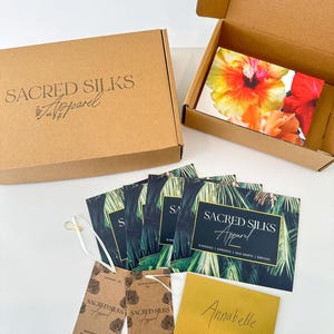 May include: Two brown cardboard boxes, one open and one closed, with the text Sacred Silks Apparel. Included are several rectangular cards featuring palm leaf patterns and the same text, two brown hang tags, and a gold envelope labeled Annabelle.