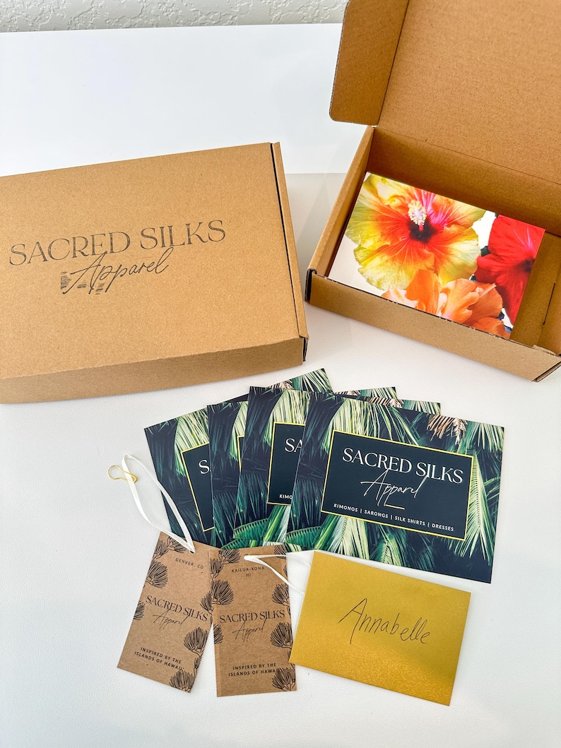 May include: Two brown cardboard boxes, one open and one closed, with the text Sacred Silks Apparel. Included are several rectangular cards featuring palm leaf patterns and the same text, two brown hang tags, and a gold envelope labeled Annabelle.