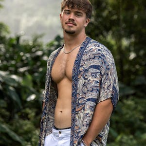 May include: A lightweight, open-front kimono-style shirt featuring a blue, beige, and white paisley pattern with a dark blue border. The garment has short, wide sleeves and a loose, flowing fit, worn over plain white trousers with a silver chain necklace.