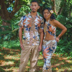 Puede incluir: Una camisa de manga corta con botones y un conjunto de dos piezas a juego que consta de un top halter y una falda larga, ambos con un estampado tropical de palmeras en blanco, naranja y azul oscuro con un patrón de borde vertical decorativo.