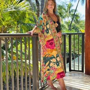 May include: A long, colorful kimono with a floral pattern in shades of pink, yellow, green, and blue. The kimono is worn over a black top. The background features lush green foliage and a wooden railing.