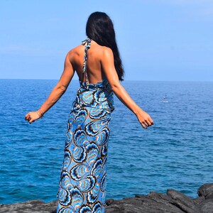 May include: A halter-neck maxi dress with a blue and white paisley pattern. The dress is worn by a person standing on a rocky shoreline, with the ocean and blue sky in the background. The dress features a flowing silhouette and a vibrant design.