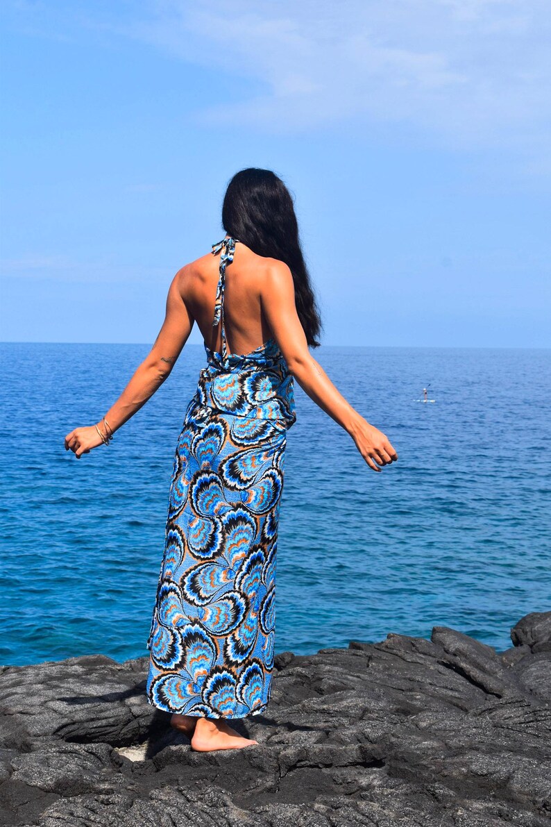 May include: A halter-neck maxi dress with a blue and white paisley pattern. The dress is worn by a person standing on a rocky shoreline, with the ocean and blue sky in the background. The dress features a flowing silhouette and a vibrant design.