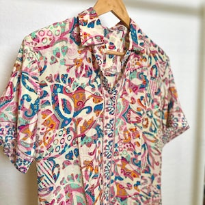 May include: Short-sleeved button-down shirt with a vibrant, colorful paisley pattern in shades of pink, blue, orange, and green on an off-white background. The shirt is hanging on a wooden hanger.