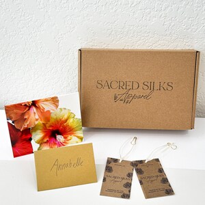May include: A brown cardboard box with the text "Sacred Silks Apparel" is next to a floral print card, a small card with the name "Anabelle", and two brown tags with the same logo. The tags are attached with white string.