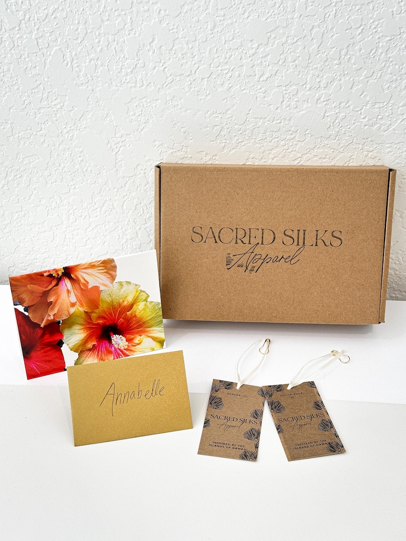 May include: A brown cardboard box with the text "Sacred Silks Apparel" is next to a floral print card, a small card with the name "Anabelle", and two brown tags with the same logo. The tags are attached with white string.