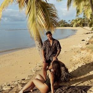 Puede incluir: Una pareja en una playa con palmeras. El hombre lleva una camisa de manga corta estampada y pantalones cortos negros. La mujer lleva un traje de ba&ntilde;o negro y est&aacute; tumbada en una manta estampada. La playa tiene arena dorada y el oc&eacute;ano est&aacute; al fondo.