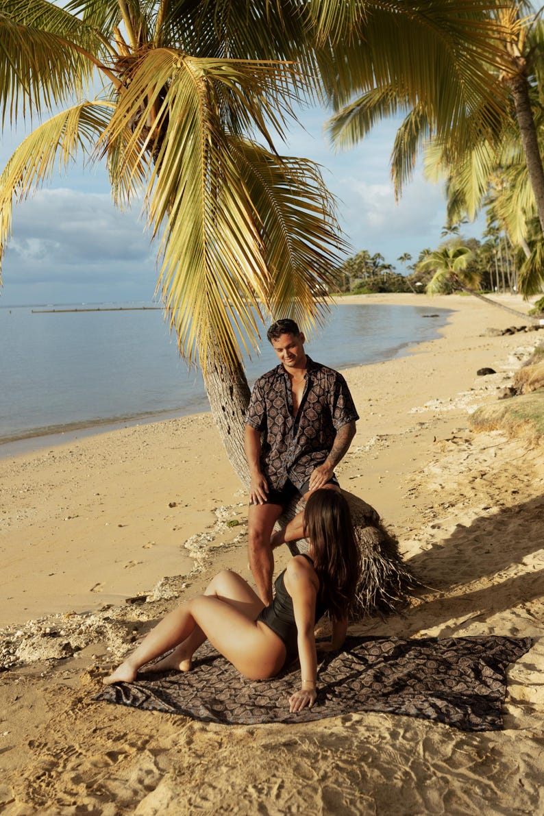 Puede incluir: Una pareja en una playa con palmeras. El hombre lleva una camisa de manga corta estampada y pantalones cortos negros. La mujer lleva un traje de ba&ntilde;o negro y est&aacute; tumbada en una manta estampada. La playa tiene arena dorada y el oc&eacute;ano est&aacute; al fondo.
