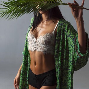 May include: A white lace bralette with a scalloped edge and a central button detail, paired with black shorts and a green kimono featuring a dark green botanical leaf pattern. The garment ensemble is worn with a palm leaf held in front of the face.