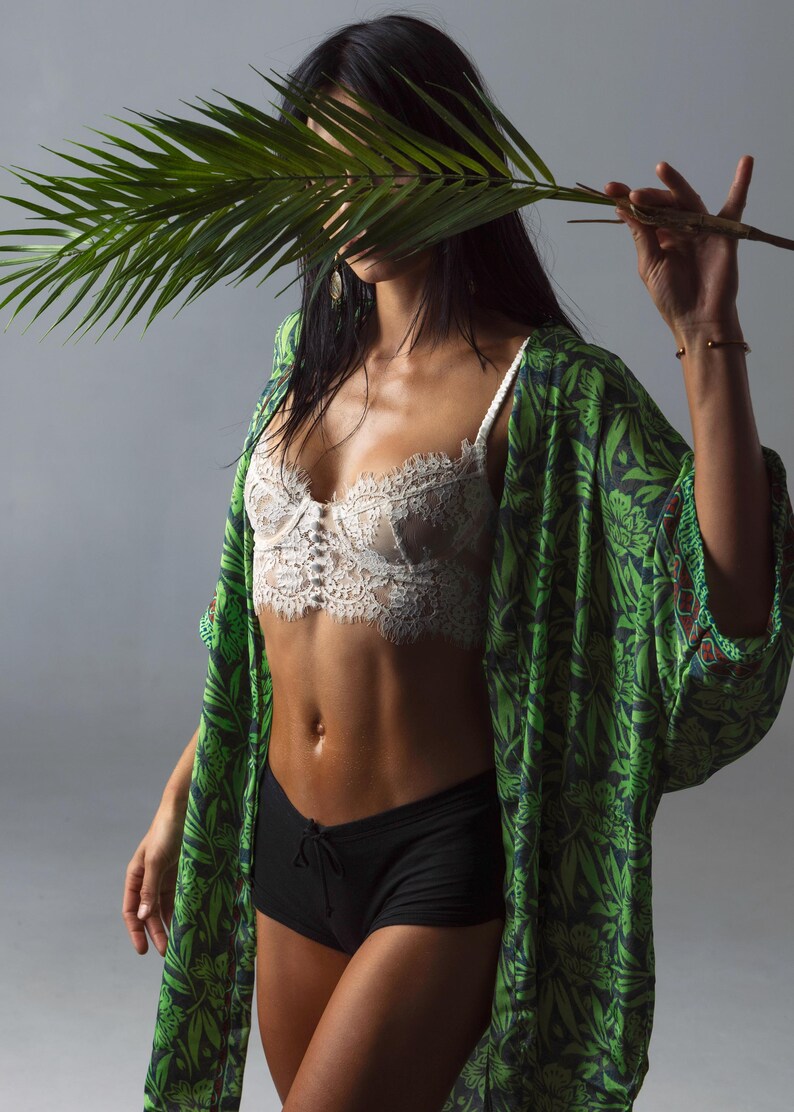 May include: A white lace bralette with a scalloped edge and a central button detail, paired with black shorts and a green kimono featuring a dark green botanical leaf pattern. The garment ensemble is worn with a palm leaf held in front of the face.
