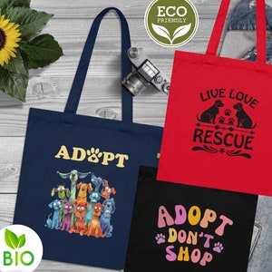 Könnte beinhalten: Drei Einkaufstaschen mit verschiedenen Designs. Die erste Tasche ist marineblau mit einer Gruppe von Cartoon-Hunden und dem Text "ADOPT". Die zweite Tasche ist rot mit dem Text "LIVE LOVE RESCUE" und einer Silhouette von drei Hunden. Die dritte Tasche ist schwarz mit dem Text "ADOPT DON'T SHOP" in bunten Buchstaben.