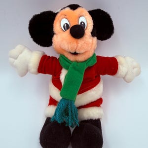 K&ouml;nnte beinhalten: Ein Stofftier Mickey Mouse, gekleidet in einer roten Jacke mit wei&szlig;em Besatz, einem gr&uuml;nen Schal und schwarzen Schuhen. Die Puppe hat gro&szlig;e schwarze Ohren, eine schwarze Nase und wei&szlig;e Handschuhe. Ein klassisches Disney-Sammlerst&uuml;ck.