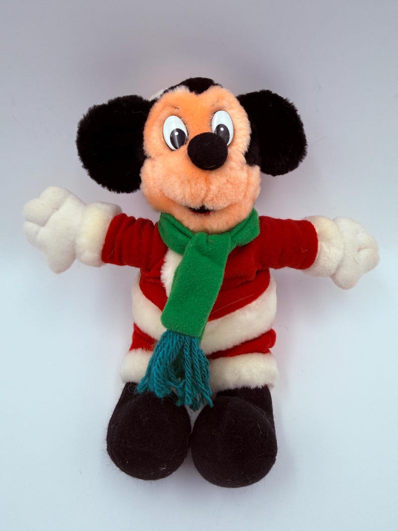 K&ouml;nnte beinhalten: Ein Stofftier Mickey Mouse, gekleidet in einer roten Jacke mit wei&szlig;em Besatz, einem gr&uuml;nen Schal und schwarzen Schuhen. Die Puppe hat gro&szlig;e schwarze Ohren, eine schwarze Nase und wei&szlig;e Handschuhe. Ein klassisches Disney-Sammlerst&uuml;ck.
