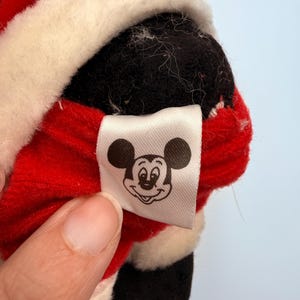 K&ouml;nnte beinhalten: Nahaufnahme eines Pl&uuml;schtiers mit einem rot-wei&szlig;en Weihnachtsmann-Outfit. Das Spielzeug hat einen schwarzen Kopf und ein wei&szlig;es Etikett mit einer Mickey Mouse-Grafik.