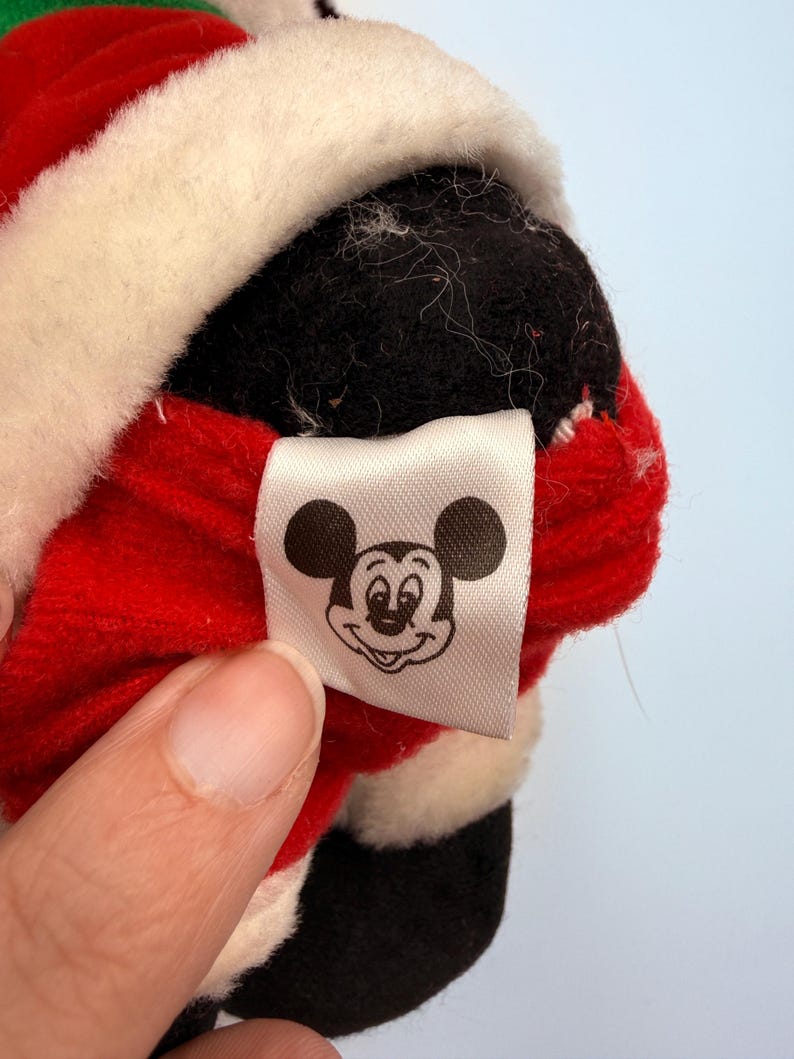K&ouml;nnte beinhalten: Nahaufnahme eines Pl&uuml;schtiers mit einem rot-wei&szlig;en Weihnachtsmann-Outfit. Das Spielzeug hat einen schwarzen Kopf und ein wei&szlig;es Etikett mit einer Mickey Mouse-Grafik.