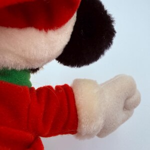 K&ouml;nnte beinhalten: Ein Pl&uuml;schtier mit roter Weihnachtsm&uuml;tze und Jacke mit wei&szlig;em Besatz. Das Spielzeug hat einen gr&uuml;nen Kragen, schwarze Ohren und einen wei&szlig;en Handschuh. Der Arm ist ausgestreckt, die Faust geballt.