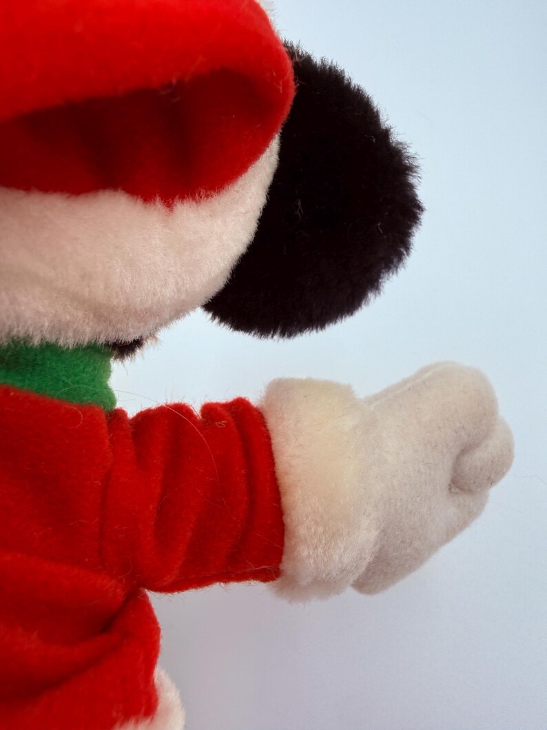 K&ouml;nnte beinhalten: Ein Pl&uuml;schtier mit roter Weihnachtsm&uuml;tze und Jacke mit wei&szlig;em Besatz. Das Spielzeug hat einen gr&uuml;nen Kragen, schwarze Ohren und einen wei&szlig;en Handschuh. Der Arm ist ausgestreckt, die Faust geballt.