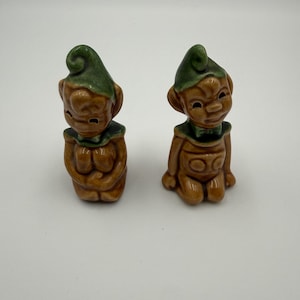May include: Two ceramic figurines of elves. Each elf has a brown body, a green hat, and a green collar. One elf is kneeling, and the other is sitting with its hands clasped.