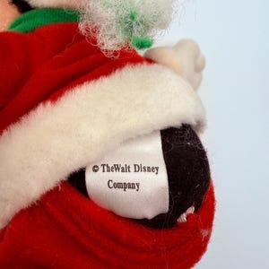 K&ouml;nnte beinhalten: Ein Pl&uuml;schtier im Weihnachtsmann-Outfit, mit einem roten Samtmantel mit wei&szlig;em Besatz und einem gr&uuml;nen Kragen. Das Spielzeug hat einen schwarzen Kopf und ein wei&szlig;es Etikett mit der Aufschrift "&copy; The Walt Disney Company."