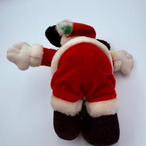 K&ouml;nnte beinhalten: Eine Pl&uuml;schpuppe des Weihnachtsmanns, von hinten gesehen, mit einem roten Anzug mit wei&szlig;er Bord&uuml;re. Die Puppe hat schwarze Schuhe, wei&szlig;e Handschuhe und einen roten Hut mit wei&szlig;er Bord&uuml;re und einem gr&uuml;nen Akzent. Auf wei&szlig;em Hintergrund.