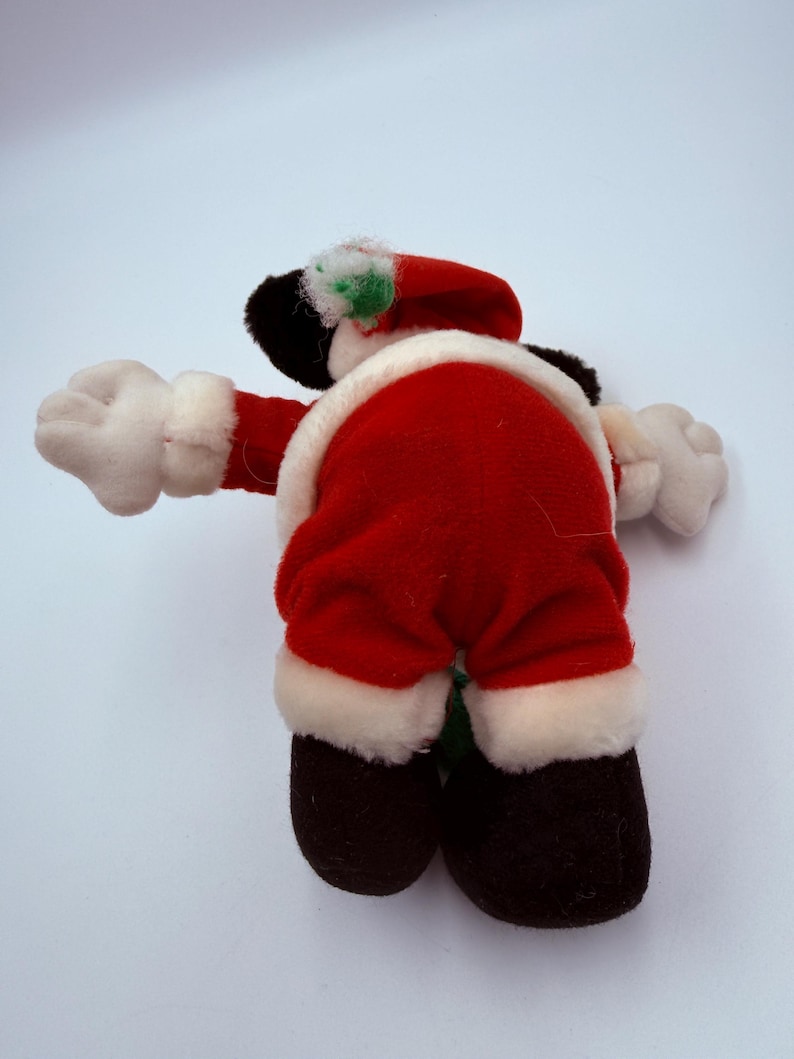 K&ouml;nnte beinhalten: Eine Pl&uuml;schpuppe des Weihnachtsmanns, von hinten gesehen, mit einem roten Anzug mit wei&szlig;er Bord&uuml;re. Die Puppe hat schwarze Schuhe, wei&szlig;e Handschuhe und einen roten Hut mit wei&szlig;er Bord&uuml;re und einem gr&uuml;nen Akzent. Auf wei&szlig;em Hintergrund.