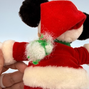 K&ouml;nnte beinhalten: Ein Pl&uuml;sch-Mickey Mouse in einem Weihnachtsmann-Outfit. Der rote Samtanzug hat einen wei&szlig;en Kunstfellbesatz. Der Hut ist rot und die Ohren sind schwarz. Ein gr&uuml;nes und wei&szlig;es Dekorationselement befindet sich um den Hals.