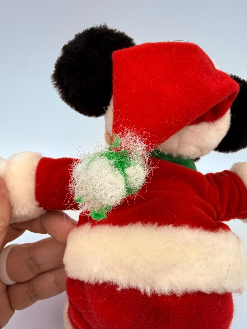 K&ouml;nnte beinhalten: Ein Pl&uuml;sch-Mickey Mouse in einem Weihnachtsmann-Outfit. Der rote Samtanzug hat einen wei&szlig;en Kunstfellbesatz. Der Hut ist rot und die Ohren sind schwarz. Ein gr&uuml;nes und wei&szlig;es Dekorationselement befindet sich um den Hals.