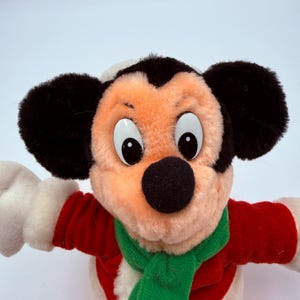 K&ouml;nnte beinhalten: Ein Stofftier von Mickey Mouse in einem festlichen Outfit. Mickey tr&auml;gt einen rot-wei&szlig; gestreiften Mantel, einen gr&uuml;nen Schal und einen wei&szlig;-schwarzen Hut. Das Spielzeug hat gro&szlig;e schwarze Ohren, eine schwarze Nase und gro&szlig;e, runde Augen.