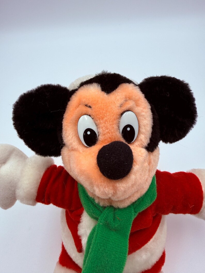 K&ouml;nnte beinhalten: Ein Stofftier von Mickey Mouse in einem festlichen Outfit. Mickey tr&auml;gt einen rot-wei&szlig; gestreiften Mantel, einen gr&uuml;nen Schal und einen wei&szlig;-schwarzen Hut. Das Spielzeug hat gro&szlig;e schwarze Ohren, eine schwarze Nase und gro&szlig;e, runde Augen.
