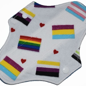 Puede incluir: Una compresa de tela blanca con varias banderas del orgullo impresas en ella, incluidas las banderas arcoíris, transgénero y bisexual. También hay pequeños corazones rojos impresos en la compresa.